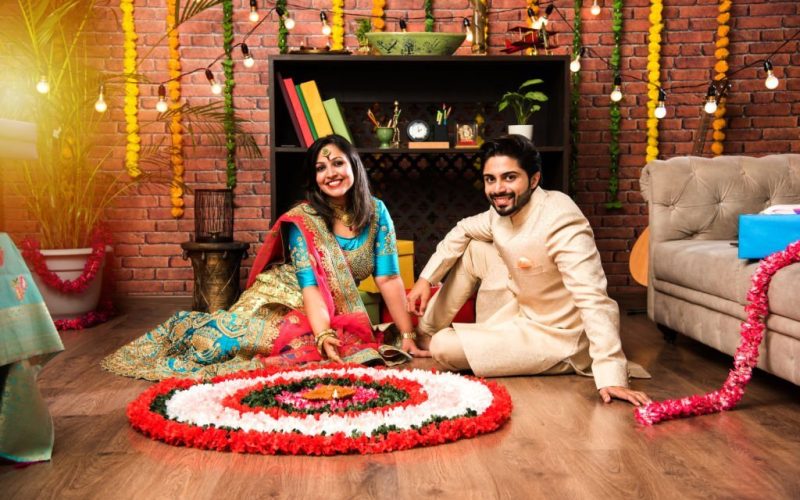 Indian couple making flower Rangoli on Diwali or Onam Festival, taking selfie or holding sweets