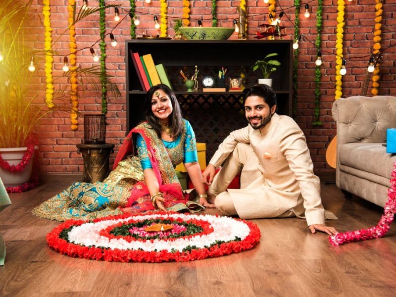 Indian couple making flower Rangoli on Diwali or Onam Festival, taking selfie or holding sweets