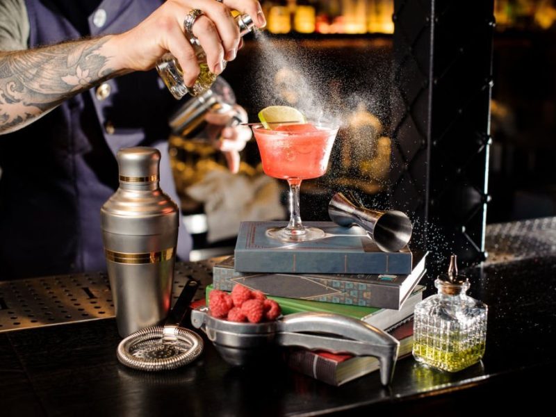 Bartender spraying on the red transparent cocktail decorated with lime with a peat bitter standing on the books on the bar counter